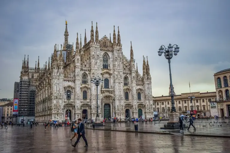 Catedral del Duomo de Milán con andamios y gente caminando por la plaza mojada bajo un cielo nublado